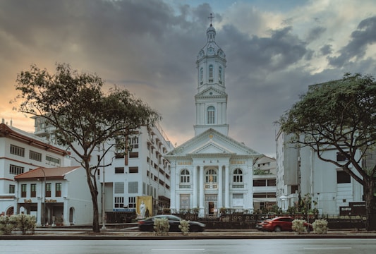 A historic church building stands prominently in the center, flanked by trees and modern urban structures. The sky above is filled with dramatic clouds at sunset, creating a rich contrast of colors.