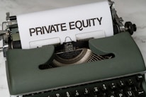 A vintage green typewriter with a sheet of paper inserted displays the text 'PRIVATE EQUITY' in bold letters. The typewriter rests on a white marble surface, and its keys and mechanisms are visible.