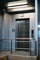 A modern elevator entrance with metallic doors and a textured floor. Safety rails are installed in front of the elevator. The area is well-lit with a fluorescent ceiling light. A wall-mounted camera is present, and there is a sign indicating wheelchair accessibility.