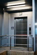 A modern elevator entrance with metallic doors and a textured floor. Safety rails are installed in front of the elevator. The area is well-lit with a fluorescent ceiling light. A wall-mounted camera is present, and there is a sign indicating wheelchair accessibility.