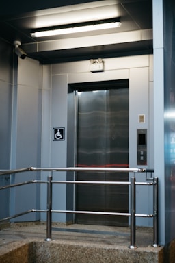 A modern elevator entrance with metallic doors and a textured floor. Safety rails are installed in front of the elevator. The area is well-lit with a fluorescent ceiling light. A wall-mounted camera is present, and there is a sign indicating wheelchair accessibility.
