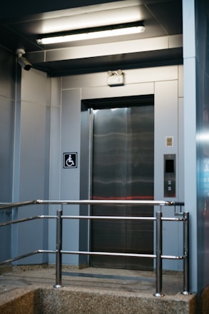 A modern elevator entrance with metallic doors and a textured floor. Safety rails are installed in front of the elevator. The area is well-lit with a fluorescent ceiling light. A wall-mounted camera is present, and there is a sign indicating wheelchair accessibility.
