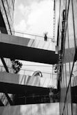 A monochromatic photograph captures a modern architectural scene, featuring a multi-story building with geometric lines and reflective surfaces. Several elevated walkways connect different parts of the structure, adorned with potted plants. The image contains stark contrasts between light and shadow, with reflections creating intricate patterns on the building surfaces. The background sky is partially visible, dotted with clouds.
