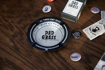A vintage-style tray with the inscription 'DAD GRASS', surrounded by various related items including buttons and packaging. The items are placed on a wooden surface, giving a rustic feel.