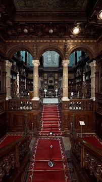 An ornate, historical library interior with intricate wooden carvings and a grand staircase covered in a red carpet. Stone columns support archways leading to the main area filled with shelves of books and statues. The ceiling features elaborate designs, and the room is dimly lit, creating a solemn atmosphere.