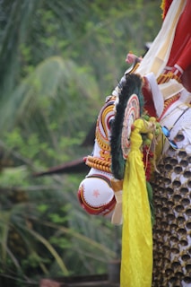 A colorful and intricately designed decorative element featuring traditional patterns and vibrant hues is prominently displayed. It includes ornate detailing with concentric circular patterns and a combination of red, yellow, black, and white colors. The background consists of lush green foliage, indicating an outdoor setting.