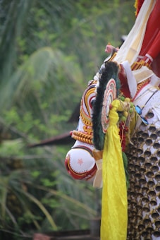 A colorful and intricately designed decorative element featuring traditional patterns and vibrant hues is prominently displayed. It includes ornate detailing with concentric circular patterns and a combination of red, yellow, black, and white colors. The background consists of lush green foliage, indicating an outdoor setting.