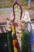 Ornate and colorful traditional Indian ornamentation featuring intricately designed faces with vibrant colors, adorned with gold embellishments and surrounded by green leaves and various colored decorations. A banner with text written in a regional script is partially visible in the background along with a person wearing a mask.