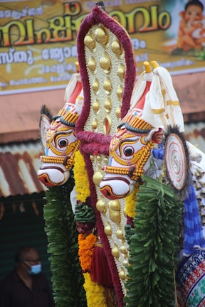 Ornate and colorful traditional Indian ornamentation featuring intricately designed faces with vibrant colors, adorned with gold embellishments and surrounded by green leaves and various colored decorations. A banner with text written in a regional script is partially visible in the background along with a person wearing a mask.