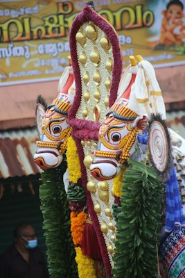 Ornate and colorful traditional Indian ornamentation featuring intricately designed faces with vibrant colors, adorned with gold embellishments and surrounded by green leaves and various colored decorations. A banner with text written in a regional script is partially visible in the background along with a person wearing a mask.
