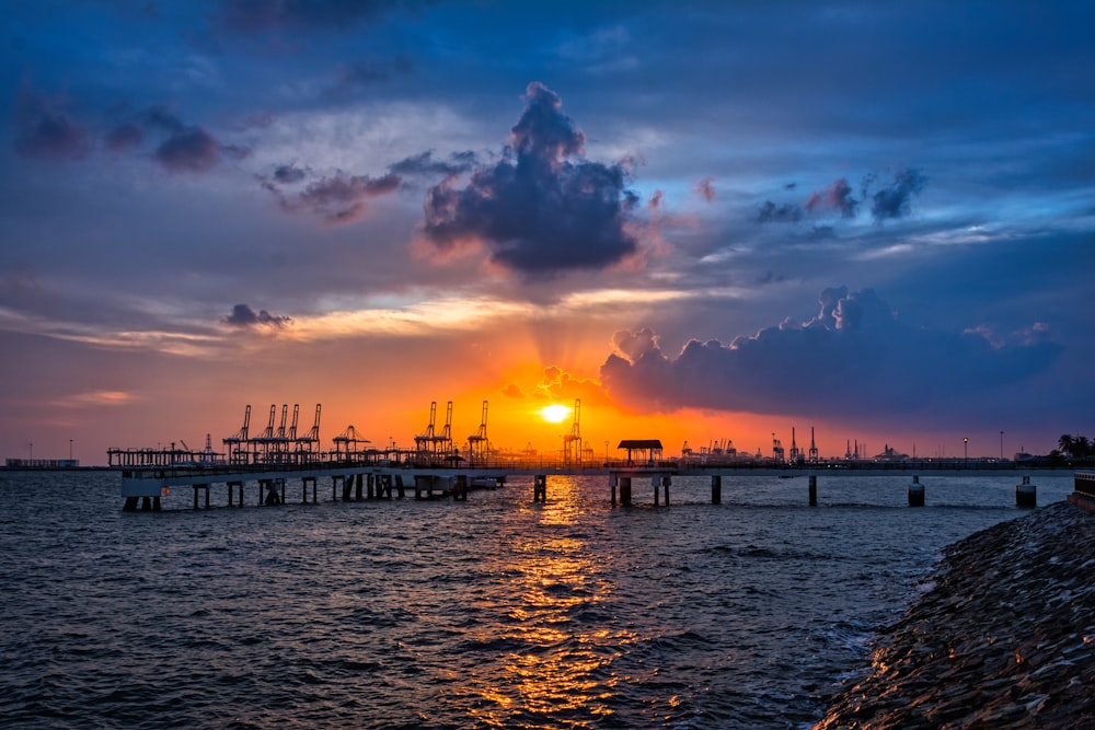 the sun is setting over the water with a pier in the foreground