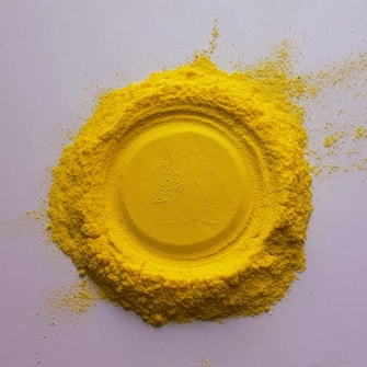 A circular pile of bright yellow powder is arranged on a flat white surface. The center is smooth, resembling an impression or mold, surrounded by looser powder scattered outward.
