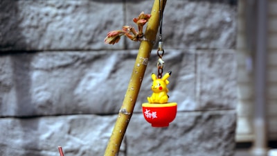 A small yellow Pikachu figurine sits inside a red cup hanging from a branch. The background features a textured, gray stone wall that is out of focus.
