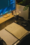A workspace features an open notebook filled with sketches and notes, placed on a dark desk surface. A large monitor displays code and applications, while a laptop with an apple logo is partially visible behind a plant. A cup with a straw sits beside the devices, indicating a working or brainstorming session.