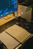 A workspace features an open notebook filled with sketches and notes, placed on a dark desk surface. A large monitor displays code and applications, while a laptop with an apple logo is partially visible behind a plant. A cup with a straw sits beside the devices, indicating a working or brainstorming session.