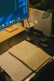 A workspace features an open notebook filled with sketches and notes, placed on a dark desk surface. A large monitor displays code and applications, while a laptop with an apple logo is partially visible behind a plant. A cup with a straw sits beside the devices, indicating a working or brainstorming session.