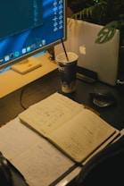 A workspace features an open notebook filled with sketches and notes, placed on a dark desk surface. A large monitor displays code and applications, while a laptop with an apple logo is partially visible behind a plant. A cup with a straw sits beside the devices, indicating a working or brainstorming session.