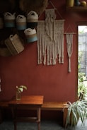 A cozy, rustic interior features a red-brown wall adorned with intricately crafted macramé wall hangings. Several woven baskets with handles hang on the wall, displaying a mix of natural and blue tones. A wooden bench and a small table occupy the space below, with a single glass bottle holding a green plant leaf. Sunlight filters through a window, accentuating the indoor greenery and decorative crafts.