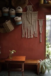 A cozy, rustic interior features a red-brown wall adorned with intricately crafted macramé wall hangings. Several woven baskets with handles hang on the wall, displaying a mix of natural and blue tones. A wooden bench and a small table occupy the space below, with a single glass bottle holding a green plant leaf. Sunlight filters through a window, accentuating the indoor greenery and decorative crafts.
