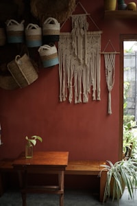 A cozy, rustic interior features a red-brown wall adorned with intricately crafted macramé wall hangings. Several woven baskets with handles hang on the wall, displaying a mix of natural and blue tones. A wooden bench and a small table occupy the space below, with a single glass bottle holding a green plant leaf. Sunlight filters through a window, accentuating the indoor greenery and decorative crafts.