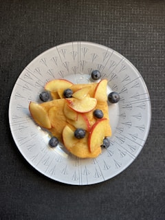 A white plate with decorative patterns contains a serving of crepes, adorned with fresh apple slices and blueberries. The plate is set against a dark textured background.