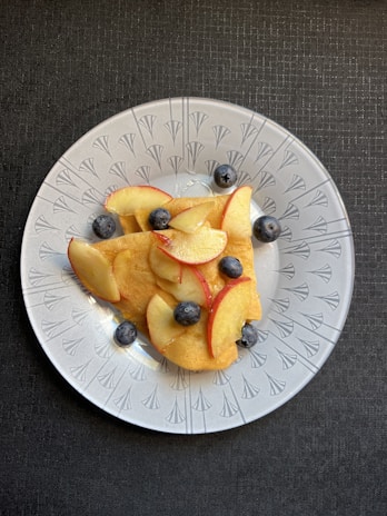 A white plate with decorative patterns contains a serving of crepes, adorned with fresh apple slices and blueberries. The plate is set against a dark textured background.