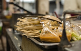A collection of skewers are placed on a metal surface alongside plastic packets containing a brownish sauce and other food items. The focus is on the skewers and the packets, with a blurred background possibly indicating a street food setting.
