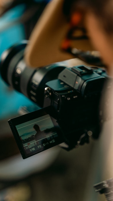 A digital camera is positioned for a shot, with its viewfinder displaying an image of a person silhouetted against a natural backdrop. The scene is focused on the camera and its screen, highlighting the camera's intricate features and the process of capturing a moment.