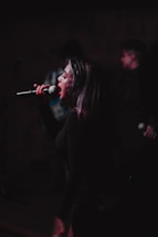 A performer passionately singing into a microphone on stage with some blurred figures in the background, suggesting a live performance or concert setting. The lighting is dim, focusing on the singer's face and expression, conveying intensity and emotion in the performance.