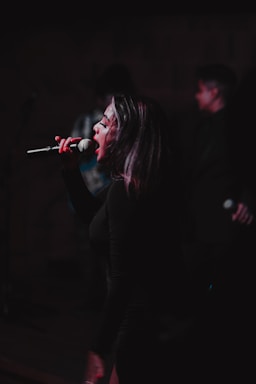A performer passionately singing into a microphone on stage with some blurred figures in the background, suggesting a live performance or concert setting. The lighting is dim, focusing on the singer's face and expression, conveying intensity and emotion in the performance.