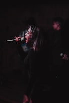 A performer passionately singing into a microphone on stage with some blurred figures in the background, suggesting a live performance or concert setting. The lighting is dim, focusing on the singer's face and expression, conveying intensity and emotion in the performance.