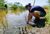 a woman crouches down to collect water from a pond
