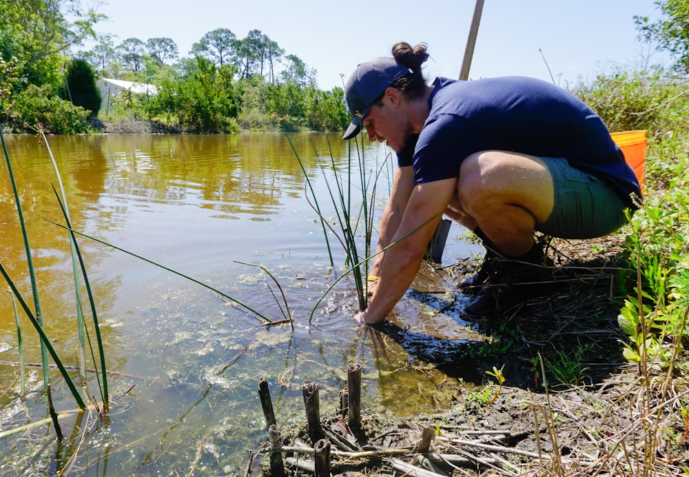 a woman crouches down to collect water from a pond