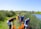A group of people are working together by a riverside, using tools and orange buckets, surrounded by tall grass and a clear blue sky.