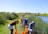 A group of people are working together by a riverside, using tools and orange buckets, surrounded by tall grass and a clear blue sky.