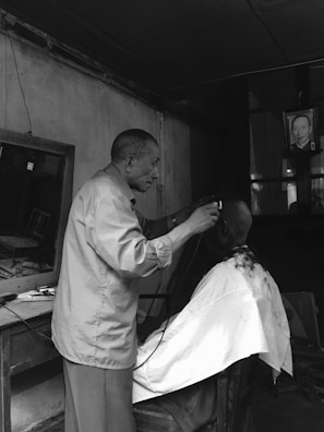 An older man is giving a haircut to another person in a small, dimly lit barbershop. The barber is using electric clippers, and both individuals appear focused on the task. The setting has an old-fashioned feel, with an aged mirror on the wall and a photograph of a person hanging nearby.