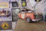 A small, vintage toy car with a weathered appearance is placed on a wooden surface. The car is predominantly red with a number '77' on its side. Surrounding the car are various artworks, including a portrait of a bearded man, an abstract painting of a person meditating, and a shelf with eclectic decorative items, including a small globe and figurines. The wall is covered with textured, colorful wallpaper featuring multiple framed pictures.