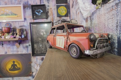 A small, vintage toy car with a weathered appearance is placed on a wooden surface. The car is predominantly red with a number '77' on its side. Surrounding the car are various artworks, including a portrait of a bearded man, an abstract painting of a person meditating, and a shelf with eclectic decorative items, including a small globe and figurines. The wall is covered with textured, colorful wallpaper featuring multiple framed pictures.