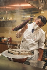 A chef wearing a white uniform and face mask is skillfully handling a piece of fish with smoke rising from a grill. The setting appears to be a professional kitchen with muted warm tones and various kitchen utensils in the background.