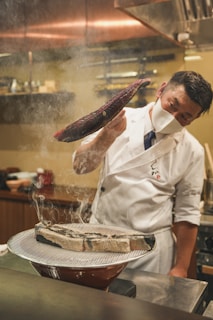 A chef wearing a white uniform and face mask is skillfully handling a piece of fish with smoke rising from a grill. The setting appears to be a professional kitchen with muted warm tones and various kitchen utensils in the background.