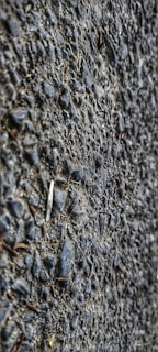 A close-up view of a textured asphalt surface with gravel. The surface is composed of small stones and coarse material, giving a rough appearance.