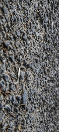 A close-up view of a textured asphalt surface with gravel. The surface is composed of small stones and coarse material, giving a rough appearance.