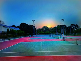 A tennis court is depicted with vibrant red and green surfaces under the early morning or evening sky. Tall floodlights and trees surround the court, and a seating area is visible in the distance. The sun is partially visible, creating a serene atmosphere.