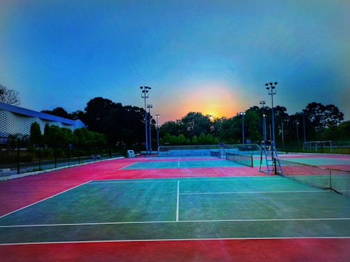 A tennis court is depicted with vibrant red and green surfaces under the early morning or evening sky. Tall floodlights and trees surround the court, and a seating area is visible in the distance. The sun is partially visible, creating a serene atmosphere.