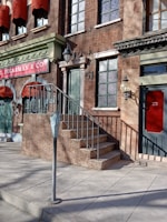 A brick building facade with red awnings and green doors, featuring a sign that reads 'Sugarman & Co Custom Furnishings Supplies'. A set of stairs with a metal railing leads up to one of the doors. A parking meter is positioned on the sidewalk in front of the building.