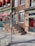A brick building facade with red awnings and green doors, featuring a sign that reads 'Sugarman & Co Custom Furnishings Supplies'. A set of stairs with a metal railing leads up to one of the doors. A parking meter is positioned on the sidewalk in front of the building.