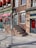 A brick building facade with red awnings and green doors, featuring a sign that reads 'Sugarman & Co Custom Furnishings Supplies'. A set of stairs with a metal railing leads up to one of the doors. A parking meter is positioned on the sidewalk in front of the building.