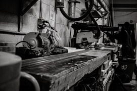 A workshop scene featuring industrial tools and machinery, including a saw on a workbench. In the background, a person, possibly a worker, is partially visible among various equipment. The setting is utilitarian and functional, with cables and metal pipes visible along the walls, contributing to a sense of busyness and productivity.