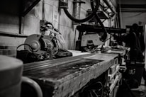 A workshop scene featuring industrial tools and machinery, including a saw on a workbench. In the background, a person, possibly a worker, is partially visible among various equipment. The setting is utilitarian and functional, with cables and metal pipes visible along the walls, contributing to a sense of busyness and productivity.