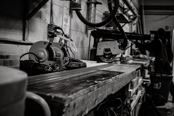 A workshop scene featuring industrial tools and machinery, including a saw on a workbench. In the background, a person, possibly a worker, is partially visible among various equipment. The setting is utilitarian and functional, with cables and metal pipes visible along the walls, contributing to a sense of busyness and productivity.